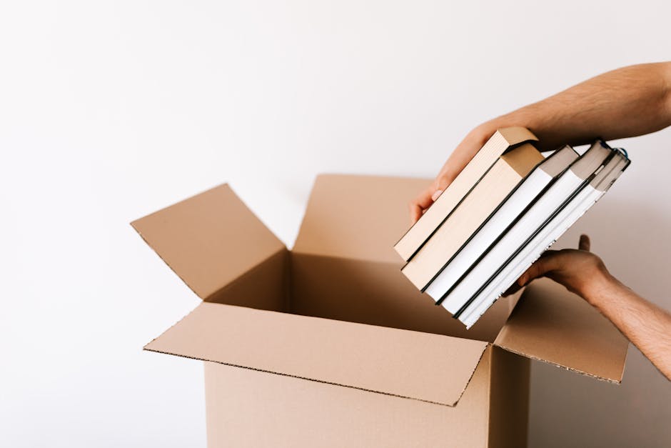 A person is seen placing a small stack of six hardcover books into an open cardboard box, with the hand supporting the bottom and the other gripping the books at the top. The box is positioned on a flat surface, likely preparing for a home relocation or packing process. The books have white and black covers and are tightly held together, ready for secure packing. The background is plain and white, emphasizing the packing activity. Additional packing materials or furniture are not visible, focusing solely on the task of organizing books for moving. This scene illustrates the packing stage of furniture transport or house removals, performed with care to ensure items are safely packed for transport, and is associated with professional moving services such as those provided by Man with Van Dagenham.