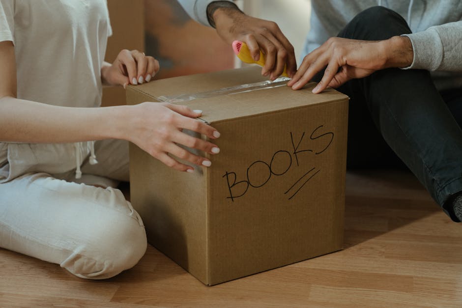 Close-up of two individuals packing a cardboard box labeled 'BOOKS' for a home relocation, with one person securing clear packing tape around the box and the other holding it steady on a wooden floor. Both are seated on the floor inside a house, with the person on the left wearing light-colored clothes and the one on the right wearing a gray long-sleeved top. The box is made of plain brown cardboard, and the word 'BOOKS' is handwritten in black marker on the side. The scene depicts the packing process associated with furniture transport and moving logistics, reflecting the careful handling of household items during a removal or house moving service, typical of operations by Man with Van Dagenham.