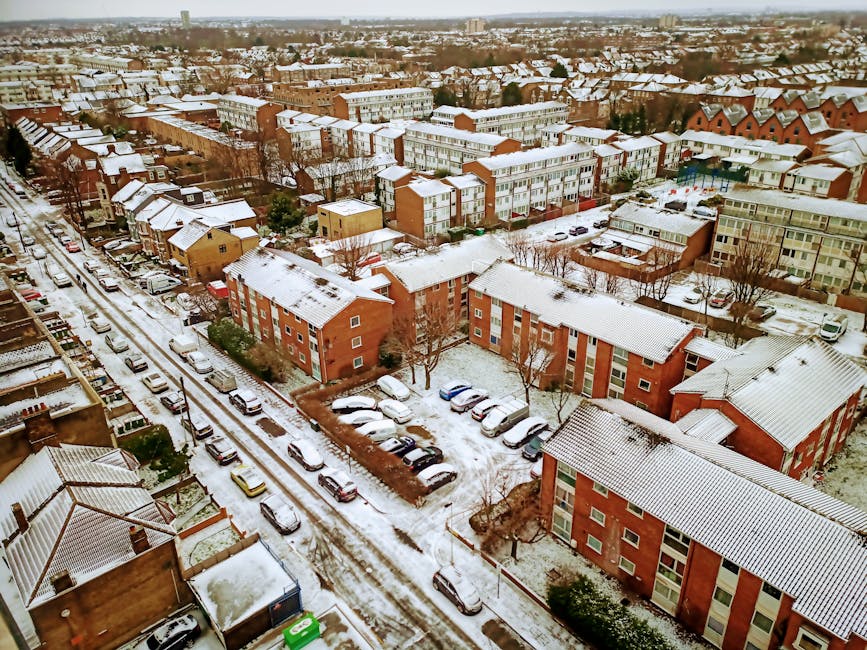 An aerial view of a residential neighbourhood during winter, showing snow-covered rooftops, streets, and parked cars. The scene features a mixture of multi-storey apartment buildings and smaller houses, with snow accumulated on the rooftops, driveways, and parked vehicles. The streets are lined with cars parked along the kerb, with tire tracks visible on the snow-covered road. The image captures the setting in Dagenham, with various brick and painted façades, leafless trees, and a broad view of the surrounding housing estate. This visual portrays a typical home relocation environment where furniture and boxes might be loaded onto delivery vans or removal trucks in preparation for moving services, as provided by Man with Van Dagenham. The overall scene emphasizes the logistics of packing, transport, and moving within an urban residential area during cold weather conditions.
