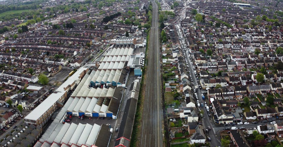 An aerial view showing a row of industrial warehouses with metal roofs split into sections, situated adjacent to a railway line running parallel through a residential area with densely packed houses, gardens, and narrow streets on either side. The warehouses are positioned on the left side of the image, with trucks and loading bays visible near the entrances. The railway line, running vertically through the centre of the image, separates the industrial zone from the housing area, where individual homes with pitched roofs, small front gardens, and parked cars line the streets. The area is set in daylight with natural lighting illuminating the scene. This image exemplifies a typical environment for home relocation and furniture transport, highlighting the logistics involved in moving goods between industrial and residential zones, essential in comprehensive removals services provided by companies such as Man with Van Dagenham.