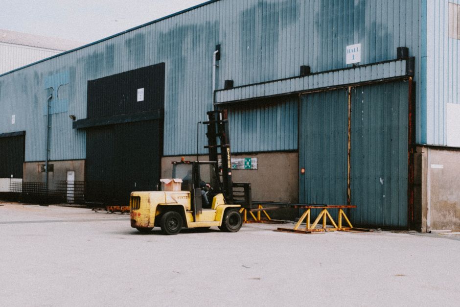 A yellow forklift truck is positioned outside a large industrial warehouse with blue corrugated metal walls, preparing for furniture transport during a home relocation or commercial move. The forklift is lifting or positioning materials near a wide, green sliding door, which is partially open, showing an interior space suitable for moving larger items like furniture and appliances. The area surrounding the warehouse is paved with concrete, with orange and yellow safety barriers placed on the ground near the forklift to facilitate safe loading operations. The scene is illuminated by natural daylight, with no active personnel in view, indicating either an idle moment in the loading process or a quiet phase of moving logistics managed by a professional removal service such as Man with Van Dagenham. Visible elements include the forklift, some packing materials or parcels on top of the vehicle, and the exterior of the warehouse, emphasizing the professional handling of business or household relocation tasks, aligned with the scope of removals services provided by [COMPANY_NAME].
