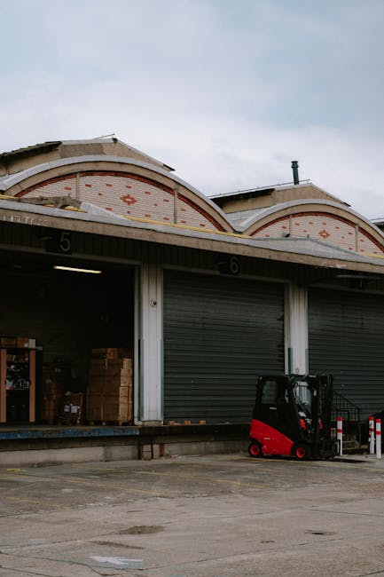 Exterior view of a warehouse with two roller shutter doors numbered 5 and 6, partially open revealing stacked cardboard boxes inside. Outside on the concrete pavement, there is a small red and black forklift positioned near the open door, ready for loading or unloading. The warehouse has a distinctive arched metal roof with decorative red and orange diamond patterns on a brick and metal façade. Overcast sky overhead creates diffuse lighting, and safety bollards are placed in front of the shutter doors. This image illustrates a typical scene from a home relocation or business removal process, where furniture and boxed items are prepared for transportation by Man with Van Dagenham as part of professional removals services for local warehouses and commercial properties.