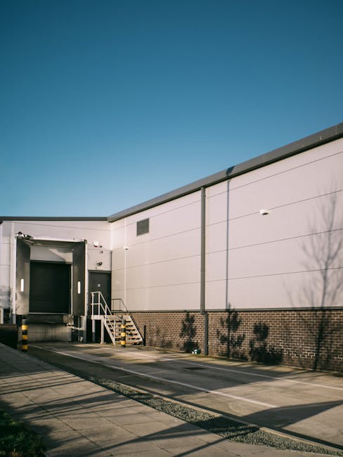 A yellow forklift truck is positioned outside a large industrial warehouse with blue corrugated metal walls, preparing for furniture transport during a home relocation or commercial move. The forklift is lifting or positioning materials near a wide, green sliding door, which is partially open, showing an interior space suitable for moving larger items like furniture and appliances. The area surrounding the warehouse is paved with concrete, with orange and yellow safety barriers placed on the ground near the forklift to facilitate safe loading operations. The scene is illuminated by natural daylight, with no active personnel in view, indicating either an idle moment in the loading process or a quiet phase of moving logistics managed by a professional removal service such as Man with Van Dagenham. Visible elements include the forklift, some packing materials or parcels on top of the vehicle, and the exterior of the warehouse, emphasizing the professional handling of business or household relocation tasks, aligned with the scope of removals services provided by [COMPANY_NAME].